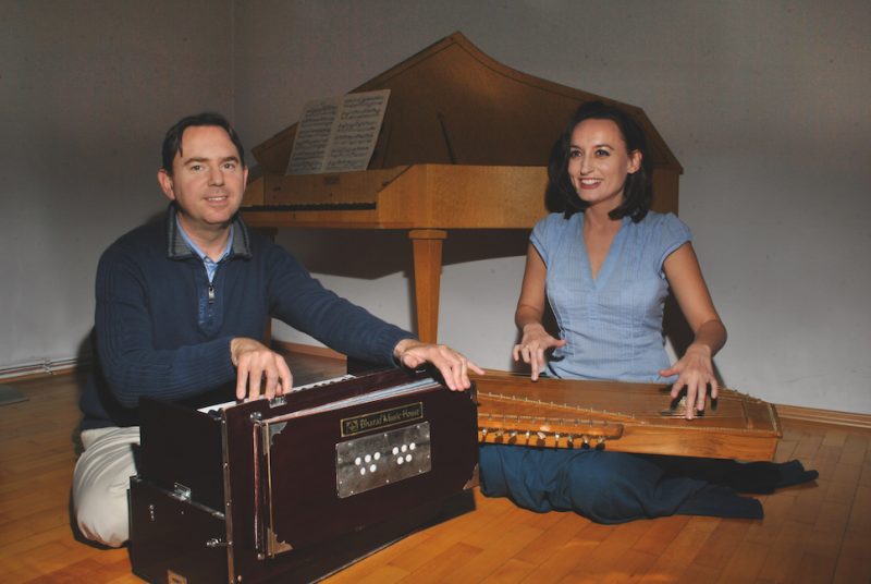 Emanuel Schulz sitzt links und spielt ein indisches Harmonium, rechts neben ihm sitzt Elisabeth Schulz und spielt Tanpura - indische Harfe. Sie sitzen am Boden und spielen heilsame Musik. Hinter ihnen ist ein Cembalo zu sehen. Alle drei Musikinstrumente sind braun. Beide Personen sind blau gekleidet. Heilsame Wirkung der Musik.